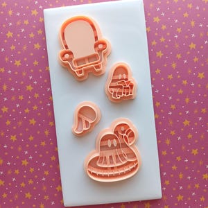 May include: A collection of peach-colored cookie cutters in various Halloween-themed shapes, including a ghost, a witch's hat, a chair, and a pair of wings. The cutters are arranged on a white rectangular surface against a pink background with gold stars.