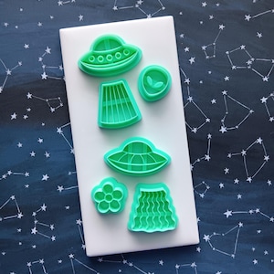May include: A set of six mint green plastic cookie cutters arranged on a white rectangular base. The shapes include two flying saucers, an alien head, a flower, and two different skirt designs with vertical and wavy line patterns.