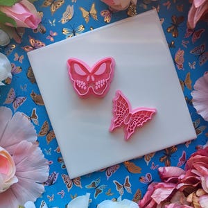 May include: Two pink plastic butterfly cookie cutters on a white surface. The surface is surrounded by a blue background with a butterfly pattern.