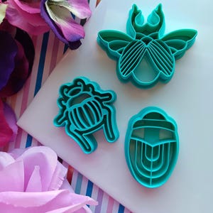 May include: Three teal blue plastic cookie cutters shaped like insects. The cutters are a beetle, a scarab beetle, and a bee.