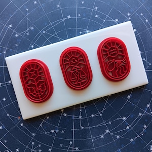 May include: Three red plastic cookie cutters with zodiac sign designs are arranged on a white rectangular surface. The cutters feature intricate details of astrological symbols. The background is a dark blue surface with a celestial map design.