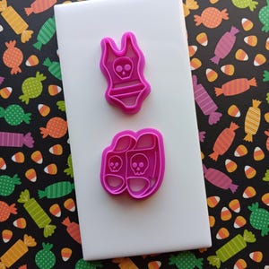 May include: Two pink plastic cookie cutters in the shape of a swimsuit and sandals, each with a skull design. The cutters are on a white surface, with a candy-themed background.