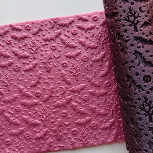 May include: A rolled and unrolled textured rolling pin with a Halloween theme. The rolling pin is dark purple and the unrolled dough is a lighter shade of purple. The pattern includes bats, skulls, trees, moons, and stars.