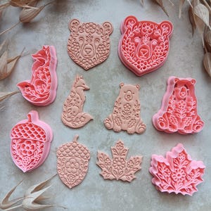 May include: A collection of ten pink plastic cookie cutters featuring intricate floral and nature-inspired designs. The set includes shapes of bears, foxes, acorns, and maple leaves, each with detailed embossed patterns for creating textured cookies.