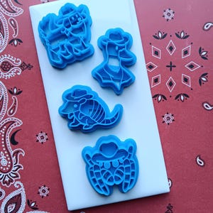 May include: Four blue plastic cookie cutters in cowboy-themed shapes: a horse, a duck, an armadillo, and a crab, all wearing cowboy hats. The cutters are arranged on a white rectangular surface, set against a red paisley patterned background.