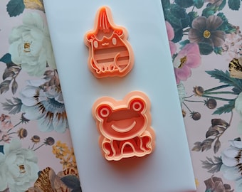 Puede incluir: Dos cortadores de galletas de plástico naranja, uno con forma de rana y el otro con forma de gato con sombrero, sobre un fondo blanco.
