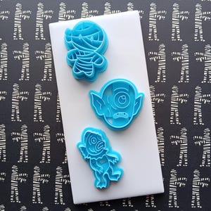 May include: Three blue plastic cookie cutters in the shapes of Halloween monsters: a mummy, a goblin, and a creature. The cutters are on a white surface, with a black and white mummy-themed background.