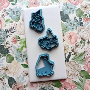 May include: Three blue plastic frog-shaped cookie cutters. Two frogs wear party hats, and one has a star. The cutters are on a white surface, set against a floral patterned background with pink and blue flowers.