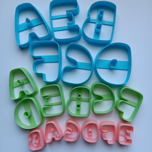 May include: A collection of plastic alphabet cookie cutters in three sizes and colors. The largest are blue, the medium are green, and the smallest are pink. Each cutter features a bold, rounded letter shape with a central support bar for stability.