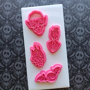 May include: A collection of pink plastic cookie cutters in various Halloween-themed shapes, including a vampire, a bat, and a devil. The cutters are arranged on a white rectangular surface.