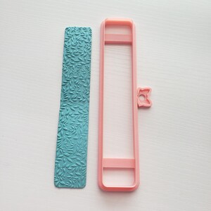 May include: A light pink plastic embossing tool with a leaf pattern and a separate frame for creating a border. The tool is designed for use with polymer clay or fondant.
