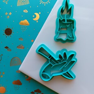 May include: Two teal-colored cookie cutters. One is shaped like a candle with a flame, and the other depicts a hand holding a smaller hand. The background is a teal surface with gold suns, moons, rainbows, and clouds.