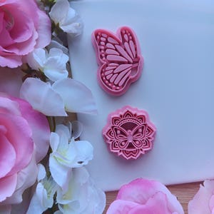 May include: Two pink butterfly-shaped cookie cutters. One is a butterfly outline, and the other is a butterfly within a floral mandala design. The cutters are on a white surface, surrounded by pink and white flowers.