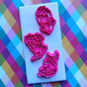 May include: Three pink plastic cookie cutters in the shapes of a woman, grapes, and a witch on a white surface. The background is a colorful plaid pattern.