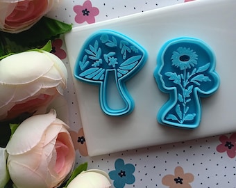 May include: Two blue plastic cookie cutters, one shaped like a mushroom with floral details and the other shaped like a person's head with a flower inside.