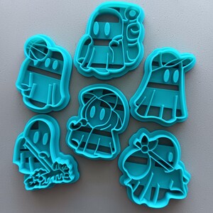 May include: Set of six turquoise blue plastic cookie cutters shaped like ghosts wearing different costumes. The ghosts are wearing a baseball cap, a raincoat, a broom, a basketball, a golf club, and a bandana.