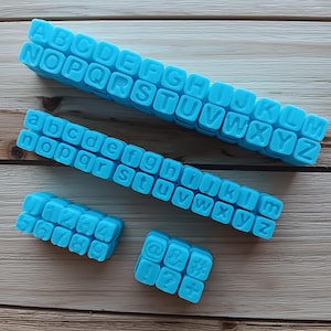 May include: A set of blue alphabet and number stamps. The stamps are arranged in rows on a wooden surface. The alphabet stamps include letters A through Z, and the number stamps include numbers 1 through 9 and 0. There are also a few special character stamps, including an at symbol, an ampersand, a hashtag, a dollar sign, a question mark, and a plus sign.