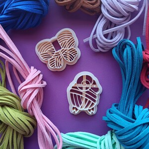 May include: An overhead shot features a collection of colorful suede cords in shades of blue, brown, pink, green, and purple. Two beige plastic cookie cutters, one butterfly-shaped and one ram-shaped, are also visible, all set against a purple background.