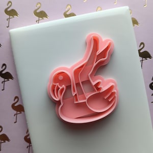 May include: A pink flamingo-shaped cookie cutter. The cutter is made of plastic and is sitting on a white surface. The background is a pink paper with gold flamingo silhouettes.
