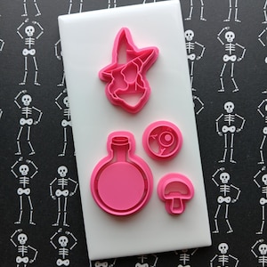 May include: Pink Halloween-themed cookie cutters on a white surface. The set includes a witch hat, potion bottle, eyeball, and mushroom shapes. The background features a black pattern with white skeleton figures.