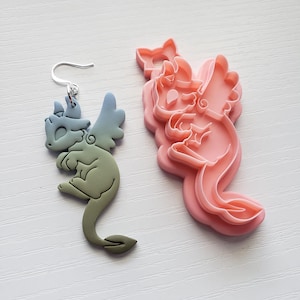 May include: A pair of blue and green dragon earrings with a silver hook. The earrings are made of polymer clay and are shaped like a dragon with wings. The earrings are shown next to a pink plastic dragon cookie cutter.