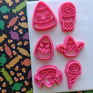 May include: A collection of six pink plastic cookie cutters in various Halloween-themed shapes. The shapes include a candy corn with a face, a brain in a basket, a candy corn with a jack-o'-lantern face, a candy corn with bat wings, a witch, and an ice cream cone.