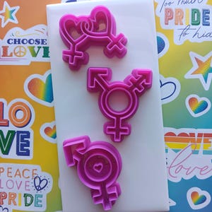 May include: Three pink plastic cookie cutters shaped like gender symbols. The first cutter is two hearts intertwined with a female symbol at the bottom. The second cutter is a transgender symbol. The third cutter is a male symbol with a heart inside the circle.