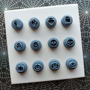 May include: A collection of twelve blue Halloween-themed cookie stamps. The stamps feature various designs, including ghosts, skulls, and pumpkins. The stamps are arranged on a white square surface against a black and silver spiderweb background.