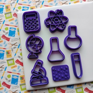 May include: A collection of purple plastic cookie cutters in various shapes, including a calculator, atom, globe, flask, microscope, and test tube. The cutters are arranged on a white surface with a colorful school-themed patterned background.