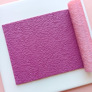 May include: A textured, magenta-colored sheet and a pink roller sit on a white surface, against a light pink background. The sheet has a detailed, cracked pattern, while the roller has a similar, but less defined texture. The items are likely for crafting or baking.