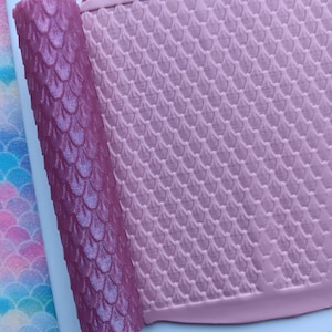 May include: A rolled-up, iridescent purple material with a scale pattern sits next to a flat, pink sheet with the same scale design. The items are on a white surface, suggesting a crafting or decorative theme.