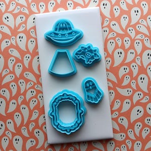 May include: A collection of blue plastic cookie cutters in various Halloween shapes, including a UFO, a ghost, and a decorative frame. The cutters are arranged on a white surface against a background of a repeating ghost pattern.