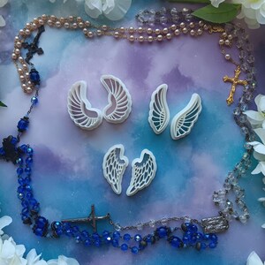 May include: Four white plastic angel wing cookie cutters on a purple and blue background with two rosaries, one with pink beads and one with blue beads.