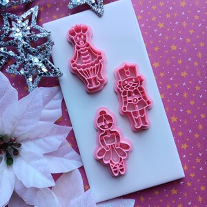 May include: Pink plastic cookie cutters in the shapes of a ballerina, a soldier, and a character with a crown. The cutters are on a white tile, with silver glitter stars and white poinsettias in the background. The background is a pink pattern with gold stars.