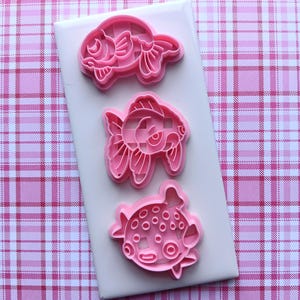 May include: Three pink plastic cookie cutters in the shape of fish are arranged on a white rectangular surface. The fish designs include a goldfish, a fish with large fins, and a pufferfish. The background is a pink and white plaid pattern.