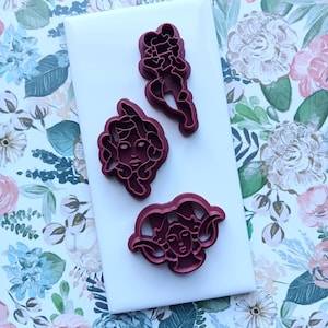 May include: Three burgundy-colored cookie cutters in various shapes, including a woman's face and a figure in motion, are arranged on a white surface. The background features a floral pattern with pink, blue, and green hues.