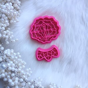 May include: Two bright pink cookie cutters. One is a detailed floral design, and the other is a smaller, decorative shape. The cutters are set against a white, fluffy background, with a string of white pearls in the corner.