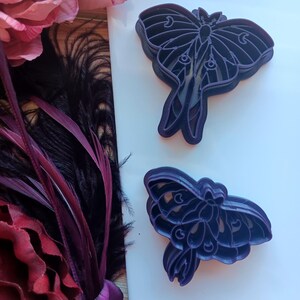 May include: Two purple plastic cookie cutters shaped like luna moths. The cutters are layered to create a 3D effect.