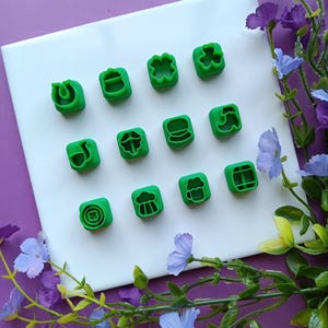 May include: A collection of green, square-shaped cookie cutters with various St. Patrick's Day-themed designs, including shamrocks and beer mugs. The cutters are arranged on a white surface, with purple and blue floral accents.