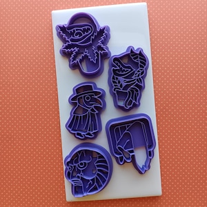 May include: A collection of five purple plastic cookie cutters in various shapes, including a plant, a creature, a plague doctor, a face, and a person in a box. The cutters are arranged on a white rectangular surface against an orange background.