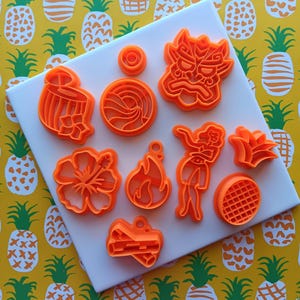 May include: A set of ten orange plastic cookie cutters shaped like various symbols and objects, including a pineapple, a hibiscus flower, a fire, a drum, a swirl, a tiki mask, a person, a leaf, and a log. The cutters are arranged on a white surface.