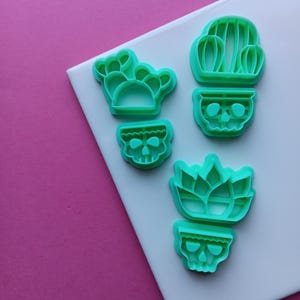May include: A set of four bright mint green plastic cookie cutters. The shapes include a multi-lobed cactus, a tall cactus in a skull-shaped pot, a small skull-shaped pot, and a succulent plant in a skull-shaped pot with defined internal details.
