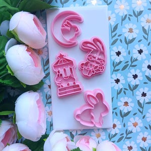 May include: Pink plastic cookie cutters in various shapes, including a bunny in a crescent moon, a bunny with a bow, a carousel, and a bunny silhouette. The cutters are arranged on a white surface, surrounded by pink flowers and a blue floral background.
