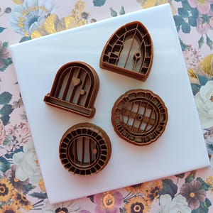 May include: Four brown plastic cookie cutters shaped like doors and windows. The cutters are on a white surface.