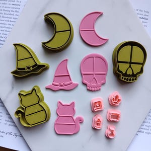 May include: A collection of plastic cookie cutters in olive green and light pink. The set includes shapes of a crescent moon, a witch hat, a skull, a cat, and four smaller items including a tombstone, a crystal, a lantern, and a cauldron.