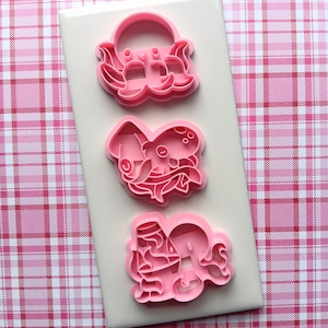 May include: Three pink plastic cookie cutters in the shape of sea creatures, including an octopus and a crab, are arranged on a white rectangular surface. The background is a pink and white plaid pattern.