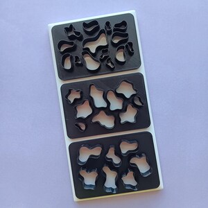 May include: Three black plastic cookie cutters with irregular shapes, arranged on a white rectangular base. The shapes resemble abstract patterns or organic forms. The cutters are likely for creating decorative cookies or pastries.