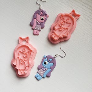 May include: Two pairs of earrings featuring a cartoon character with a single large eye, one pair is pink and the other is blue. The earrings are made of polymer clay and come with silver ear wires.