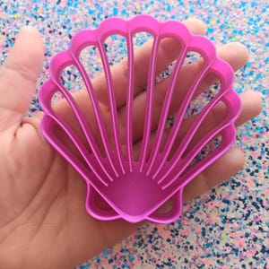 May include: A pink plastic seashell cookie cutter with a detailed design. The cutter has a scalloped edge and a raised center with multiple ridges.