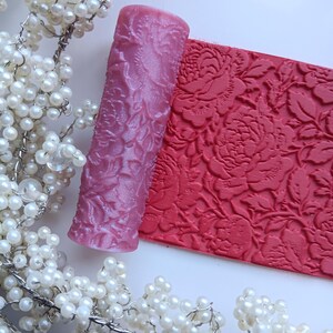 May include: A pink rolling pin and a red textured mat with a floral pattern. The rolling pin is cylindrical with a detailed embossed design. The mat features a repeating rose pattern. Decorative pearl garland frames the objects.
